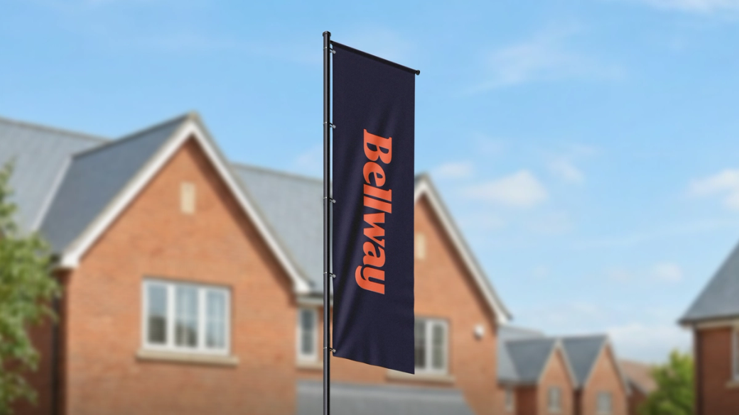 The image shows a flag displaying the name 'Belway' prominently in front of a residential area with brick houses. The setting suggests a construction or real estate context.