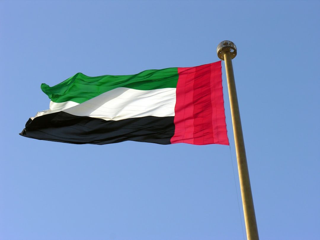 The image shows the flag of the United Arab Emirates waving against a clear blue sky. The flag features horizontal stripes of green, white, black, and red.