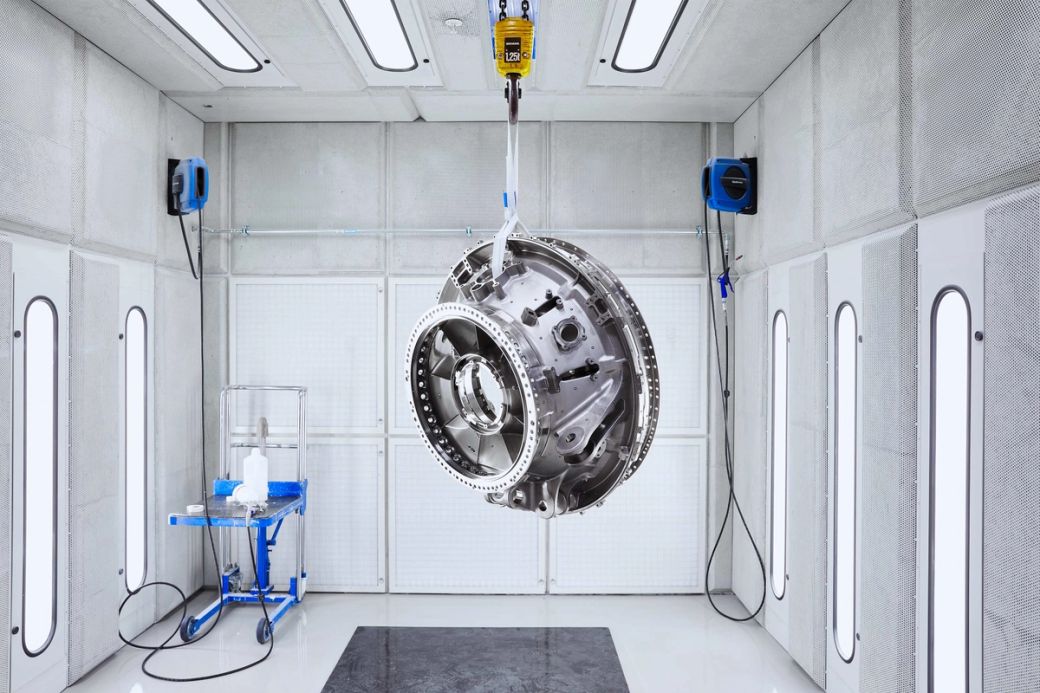 The image features a large metallic engine component suspended from a ceiling hook inside a clean, industrial workspace. The environment is well-lit and designed for maintenance or assembly of precision parts.