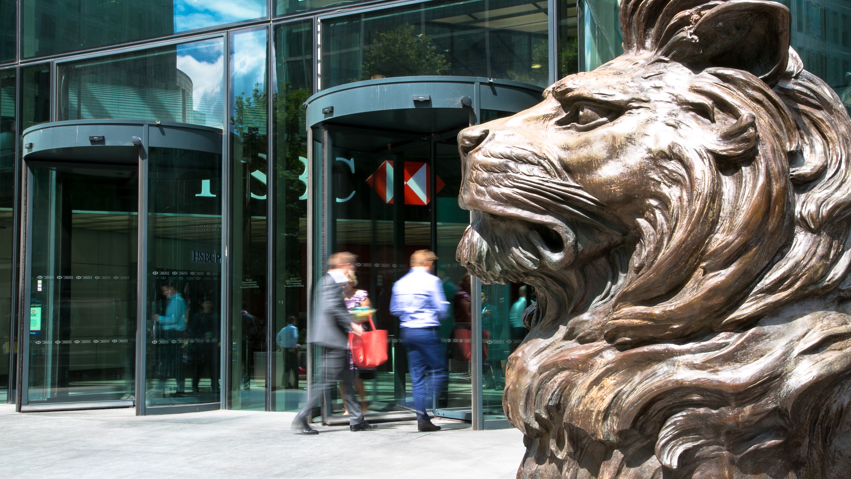The image captures the entrance of a modern corporate building, featuring a large bronze lion sculpture in the foreground. Two professionals, dressed in business attire, are walking through the revolving doors, suggesting an active business environment.