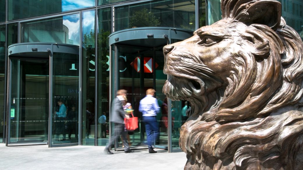 The image captures the entrance of a modern corporate building, featuring a large bronze lion sculpture in the foreground. Two professionals, dressed in business attire, are walking through the revolving doors, suggesting an active business environment.