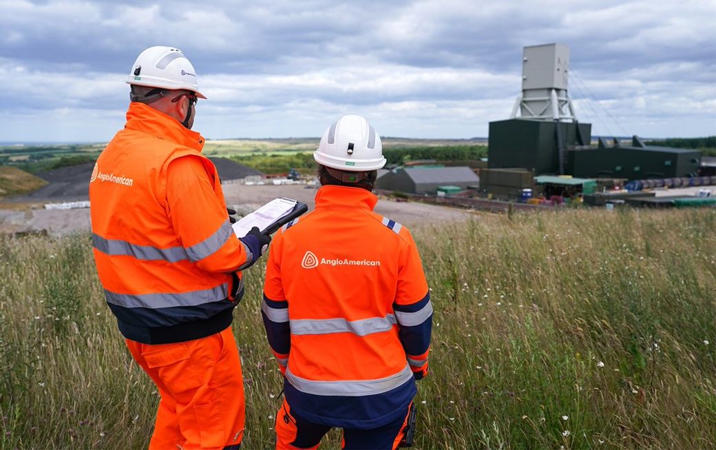 Two workers in safety attire stand on a hillside overlooking a mining site. One worker is holding a clipboard while the other is monitoring the environment, indicating active operations in a mining setting.