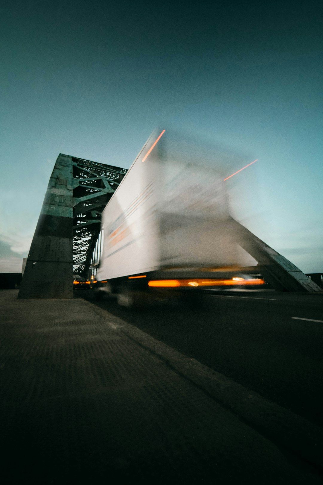 The image shows a blurred motion of a white truck crossing a bridge at dusk. The structure of the bridge is prominently visible, with the darkening sky providing a contrasting background.