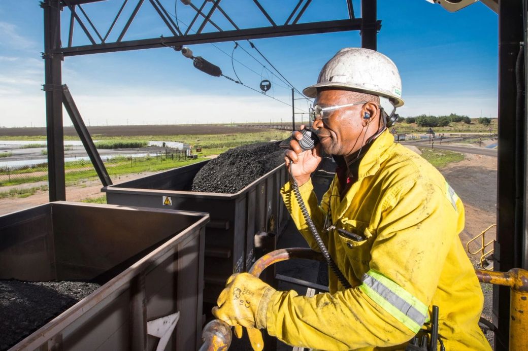 A worker in a yellow safety jacket and white hard hat stands on a railing, speaking into a radio while overseeing coal being loaded into a freight train. The background features a landscape of fields and power lines under a clear blue sky.