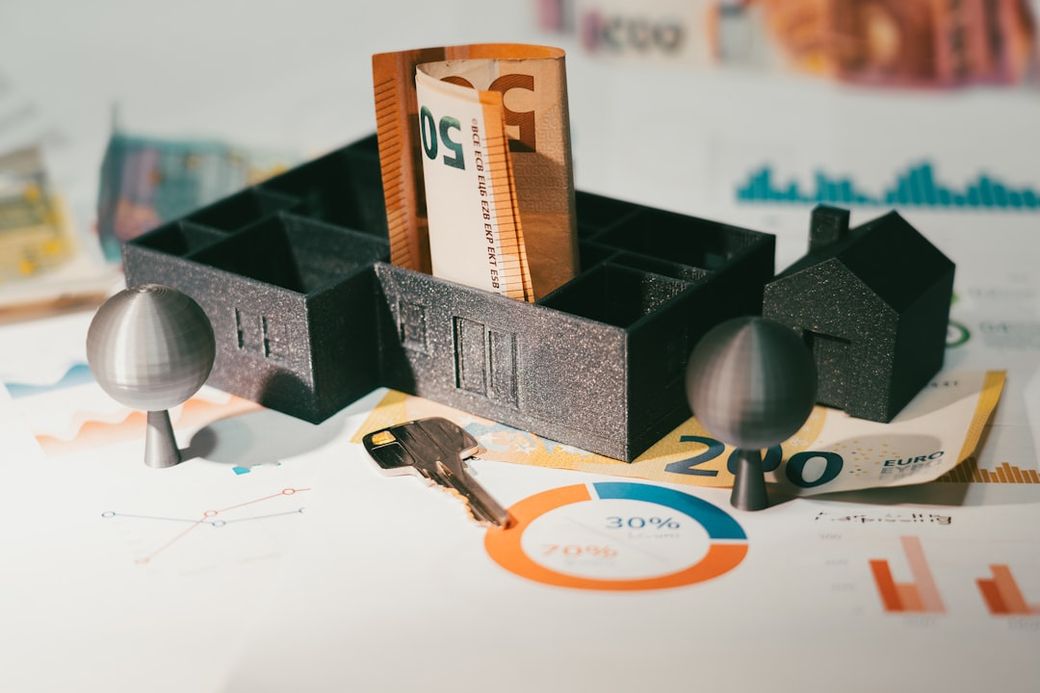 The image depicts a miniature model of a building with a rolled banknote placed prominently inside it, surrounded by various financial charts and keys. It suggests concepts related to real estate and finance. — Credit: Photo by Jakub Żerdzicki on Unsplash