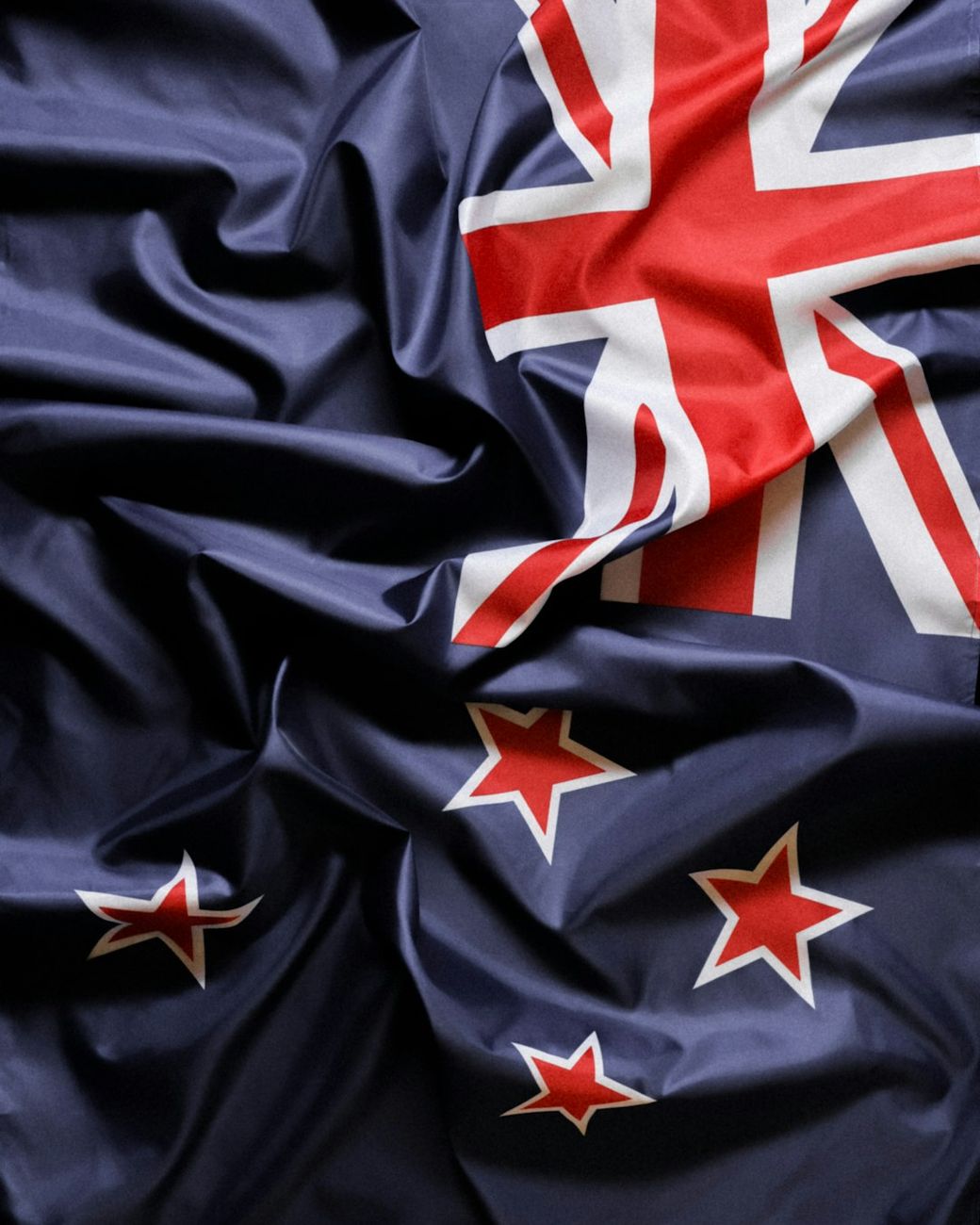 The image features a close-up of a wrinkled flag of New Zealand, showcasing its blue background, Union Jack, and Southern Cross constellation with five white stars. The fabric's texture is emphasized through the folds and creases. — Credit: Photo by Saymom Leão on Unsplash