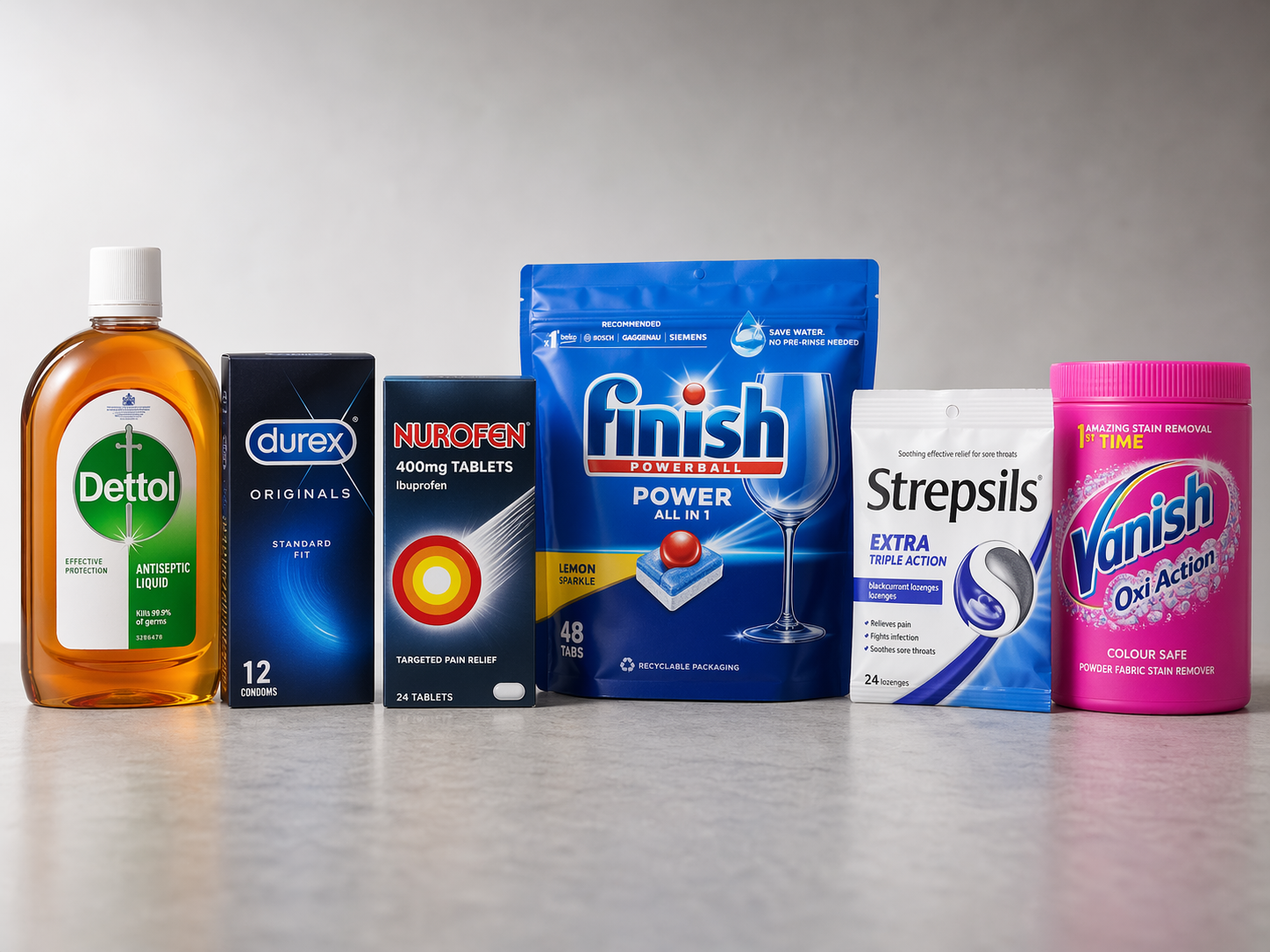 The image displays a selection of healthcare and household products arranged in a linear formation. Notable items include a disinfectant, condoms, pain relief tablets, a dishwasher detergent, throat lozenges, and a stain remover.