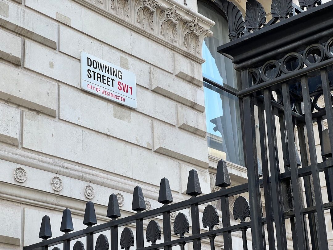 The image depicts a close-up view of a sign indicating Downing Street, located in the City of Westminster. The architectural details of the building are visible alongside a decorative iron gate.