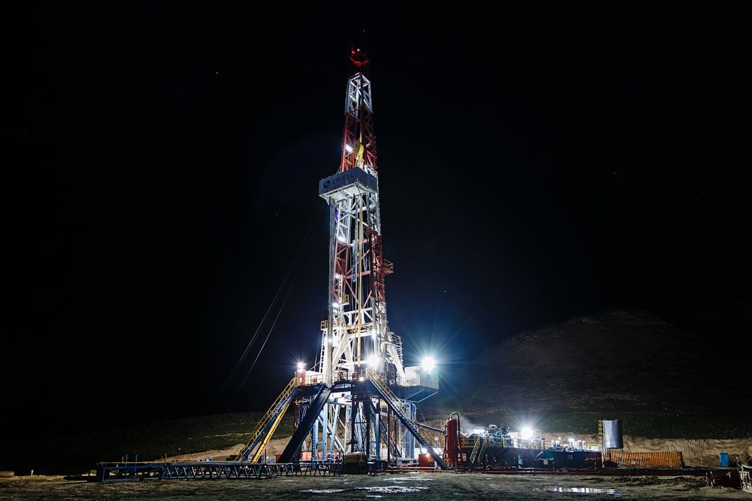 The image depicts a drilling rig illuminated at night, showcasing its structure and equipment in detail. The surrounding area is dark, emphasizing the rig's lights and machinery.