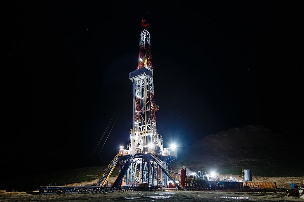 The image depicts a drilling rig illuminated at night, showcasing its structure and equipment in detail. The surrounding area is dark, emphasizing the rig's lights and machinery. — Credit: Photo by WORKSITE Ltd. on Unsplash