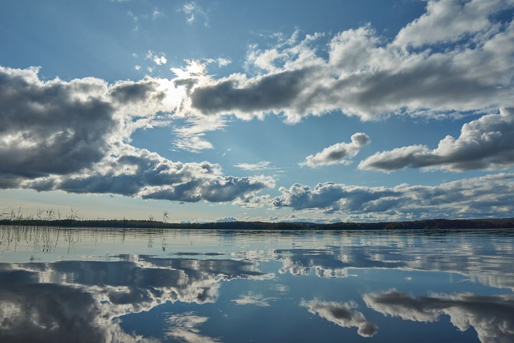 A serene landscape featuring a calm body of water reflecting a sky filled with clouds. Sunlight breaks through the clouds, creating a picturesque view of nature. — Credit: Photo by Wolfgang Hasselmann on Unsplash