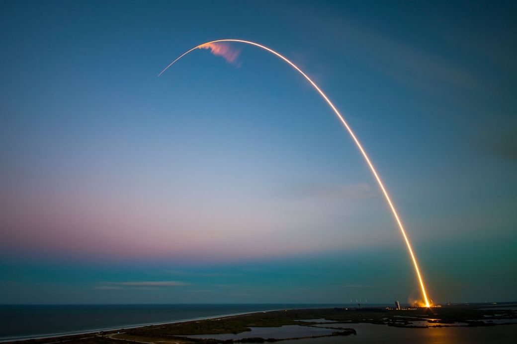 The image captures the launch trajectory of a rocket against a twilight sky, creating a bright arc of light. The scene depicts the launch site near a body of water, with the rocket's launch producing a visible plume of smoke and flames. — Credit: Photo by SpaceX on Unsplash
