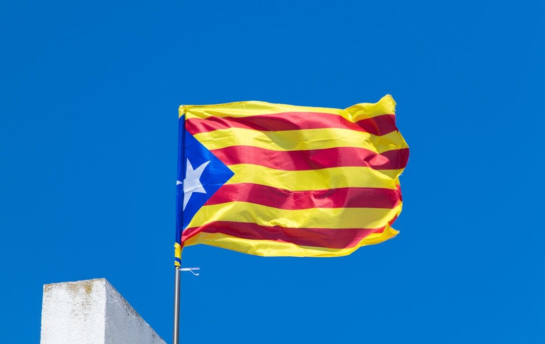 The image features the flag of Catalonia waving against a clear blue sky. The flag, known as 'Senyera', consists of red and yellow stripes with a blue triangle containing a white star.