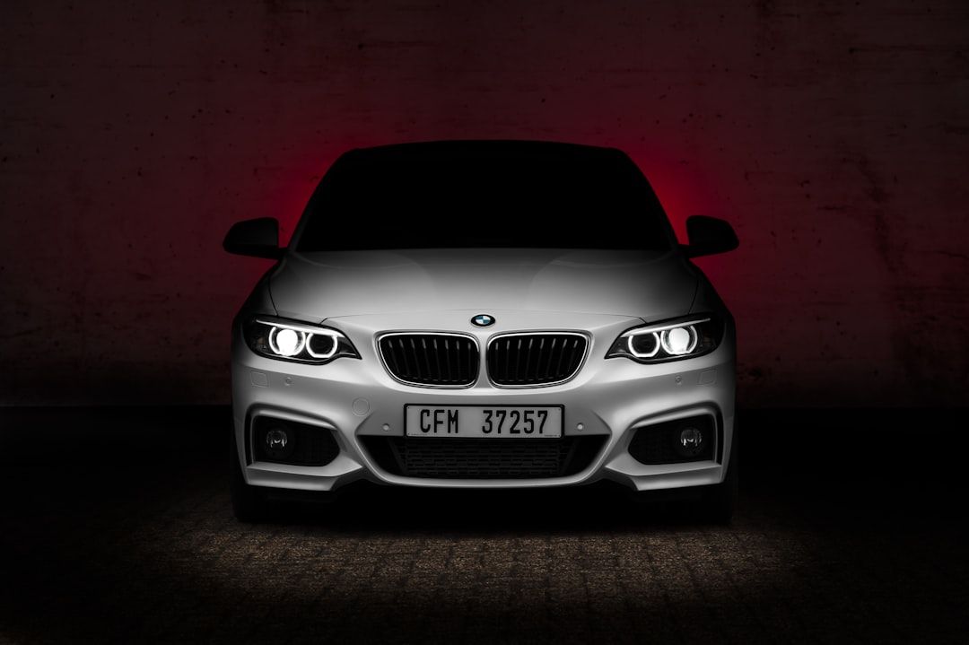 The image features a front view of a BMW car, showcased against a dark, textured background with red lighting accents. The vehicle's sleek design is highlighted by the bright headlights and distinctive kidney grille.