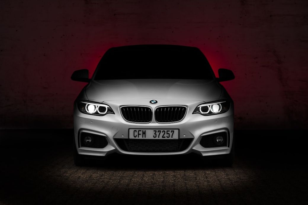The image features a front view of a BMW car, showcased against a dark, textured background with red lighting accents. The vehicle's sleek design is highlighted by the bright headlights and distinctive kidney grille. — Credit: Photo by Stephan Louis on Unsplash