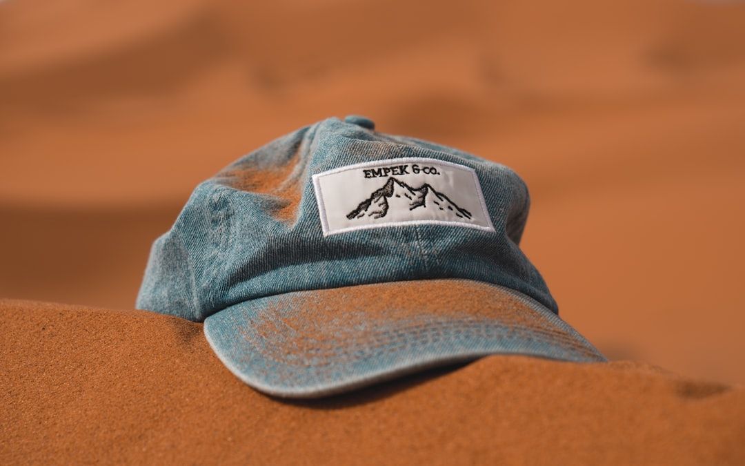 The image features a blue cap with a mountain logo positioned on a sandy surface, suggesting a desert or outdoor setting. The cap bears the brand name 'EMPEK & CO.' prominently displayed on the front.
