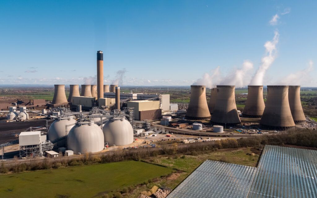 The image depicts a large power generation facility featuring cooling towers and industrial infrastructure. Steam is emitted from the towers, indicating active operations, set against a clear blue sky.