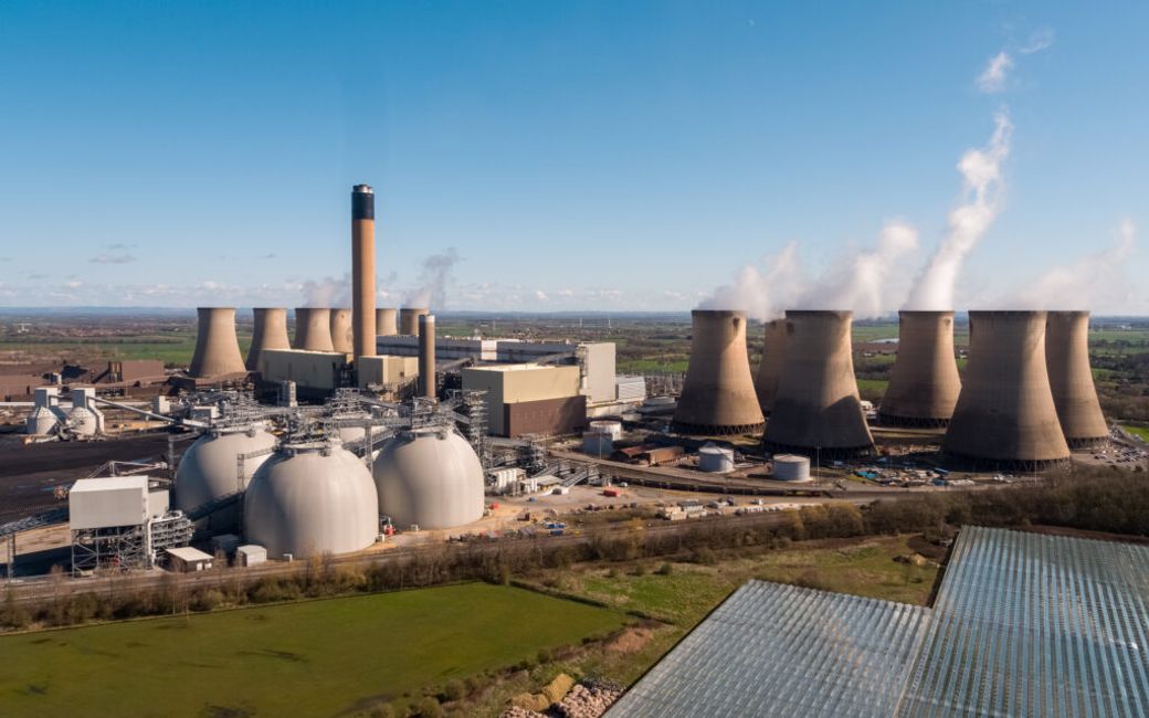 The image depicts a large power generation facility featuring cooling towers and industrial infrastructure. Steam is emitted from the towers, indicating active operations, set against a clear blue sky.