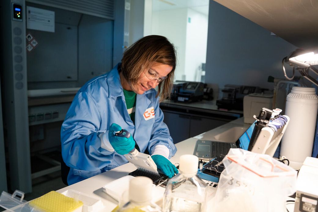A laboratory technician wearing protective gear is engaged in a scientific experiment, using a pipette to handle samples at a workstation. The setting is a modern laboratory with various equipment and tools visible in the background.