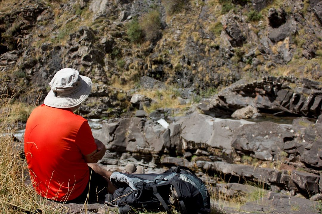 A man wearing a wide-brimmed hat sits on a rocky outcrop, gazing at a rugged landscape. He appears to be enjoying a moment of solitude in nature, surrounded by dry grass and rocky terrain.