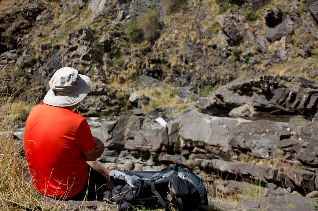 A man wearing a wide-brimmed hat sits on a rocky outcrop, gazing at a rugged landscape. He appears to be enjoying a moment of solitude in nature, surrounded by dry grass and rocky terrain. — Credit: Photo by Victor Morales Cayuela on Unsplash