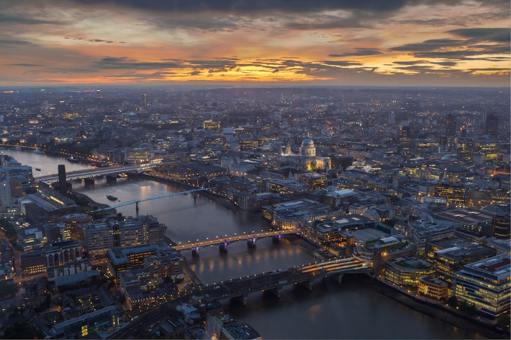 The image depicts a stunning aerial view of London during sunset, showcasing the skyline with prominent landmarks such as the River Thames and St. Paul's Cathedral. The vibrant colors of the sunset reflect off the buildings and water, creating a picturesque urban landscape. — Credit: Photo by Jaanus Jagomägi on Unsplash