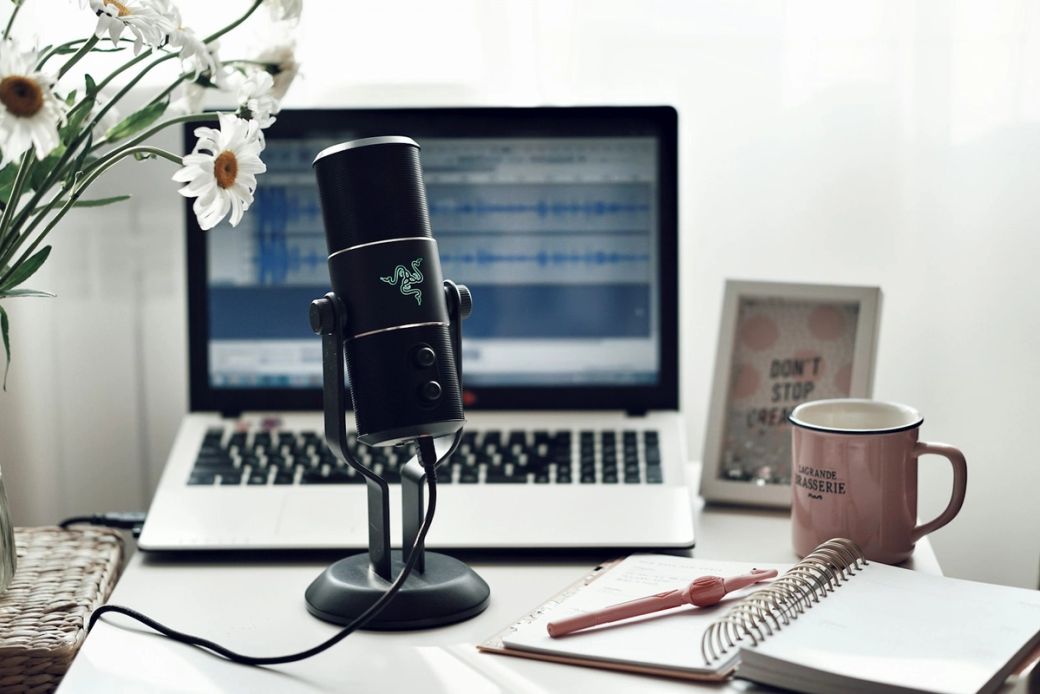 A desktop podcasting setup featuring a microphone, laptop, and a notebook. The background shows audio editing software on the laptop screen, while a coffee mug and flowers add a personal touch to the workspace. — Credit: Photo by Vika Strawberrika / Unsplash