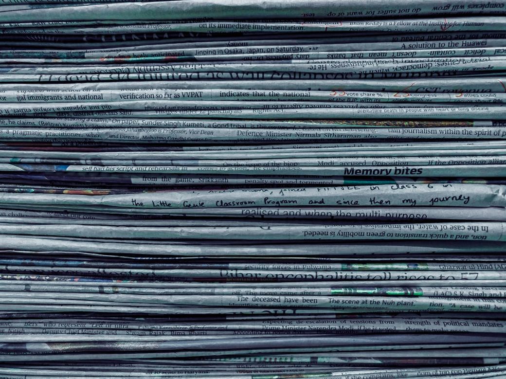 A stack of newspapers with various articles visible. The papers are printed in black and white, showcasing headlines and segments of text that suggest coverage of diverse topics. — Credit: Photo by Utsav Srestha on Unsplash