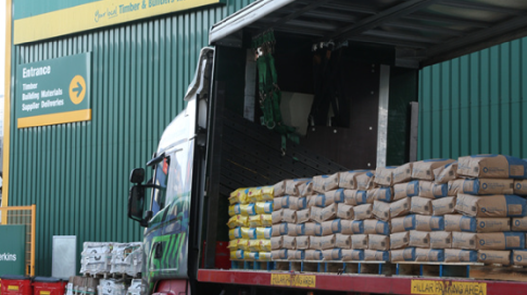 A delivery truck is parked at a construction supply facility, unloading bags of building materials. The backdrop features signage indicating the entrance and services offered at the location.