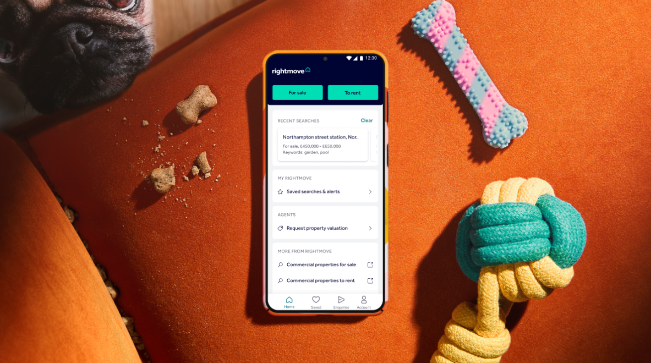 The image features a smartphone displaying the Rightmove real estate application interface. In the background, a playful dog peeks out beside a brightly colored orange cushion and several dog toys, creating a lively home setting.