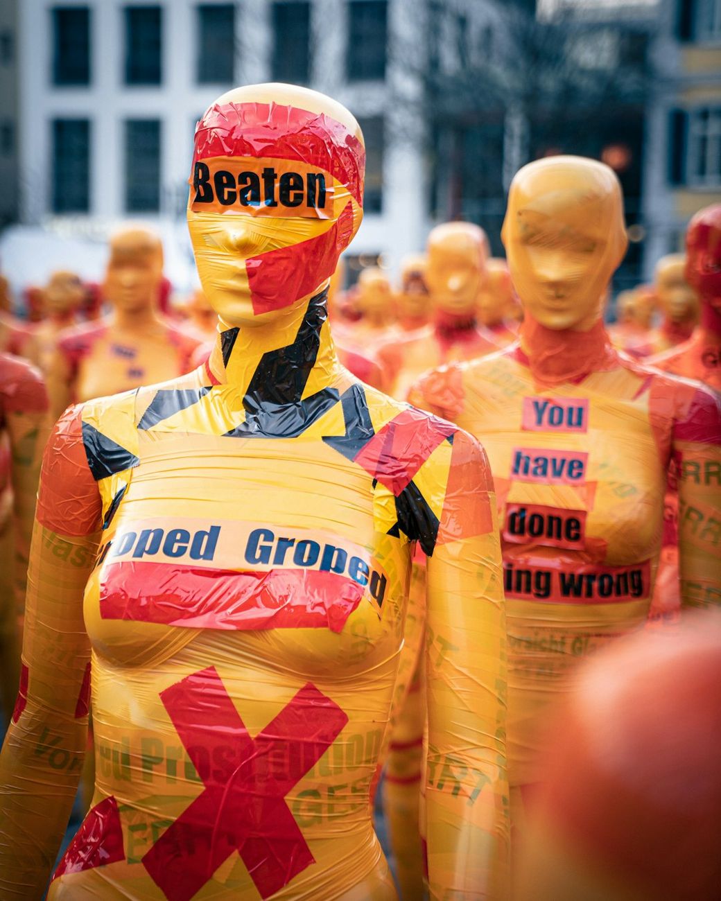 The image features a large group of mannequins covered in bright orange and yellow plastic wrap, each adorned with various phrases and symbols including 'Beaten' and 'You have done wrong.' They are arranged in a protest-like formation, indicating a message or demonstration. — Credit: Photo by Mika Baumeister on Unsplash
