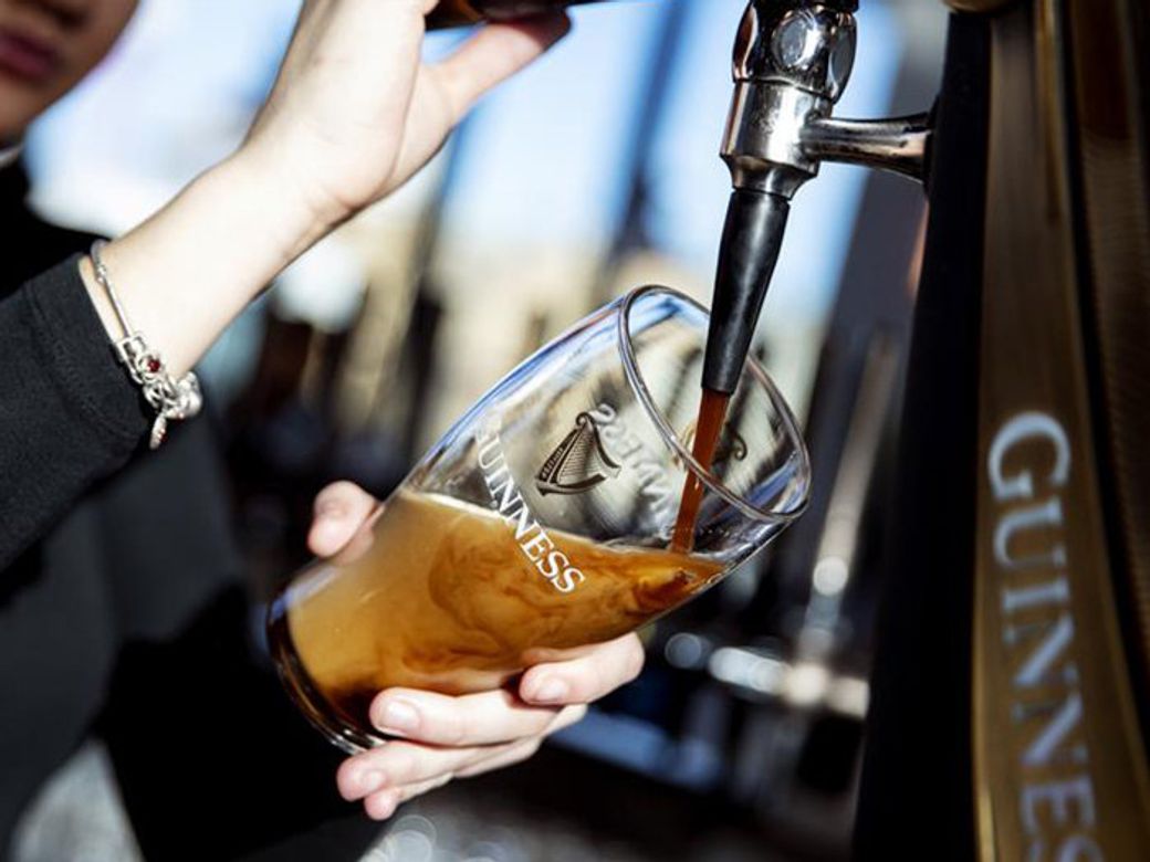 A bartender is pouring a pint of Guinness beer into a branded glass at a bar. The image captures the action of the beer flowing from the tap into the glass.