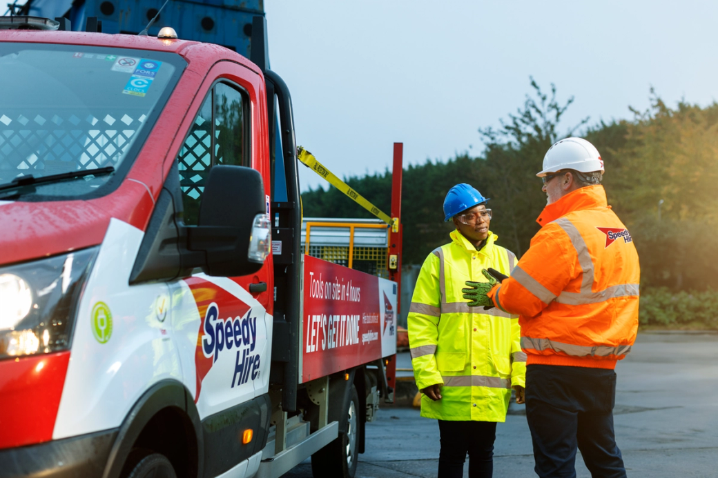 A construction site scene depicting two individuals in safety gear engaging in a conversation. One person is wearing a high-visibility yellow jacket and a hard hat, while the other is dressed in a similar style. A truck with branding is parked nearby.