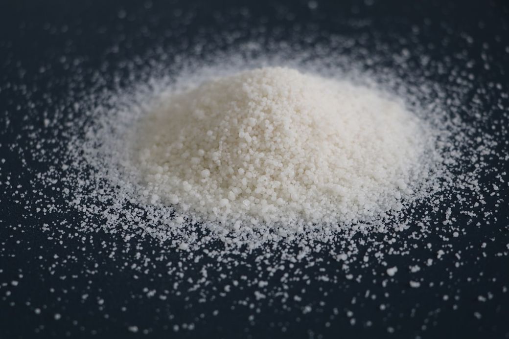 A close-up image of a pile of white granulated substance resembling sugar or salt. The texture and details of the crystalline structure are prominently visible against a dark background. — Credit: Photo by XRD Chemical on Unsplash
