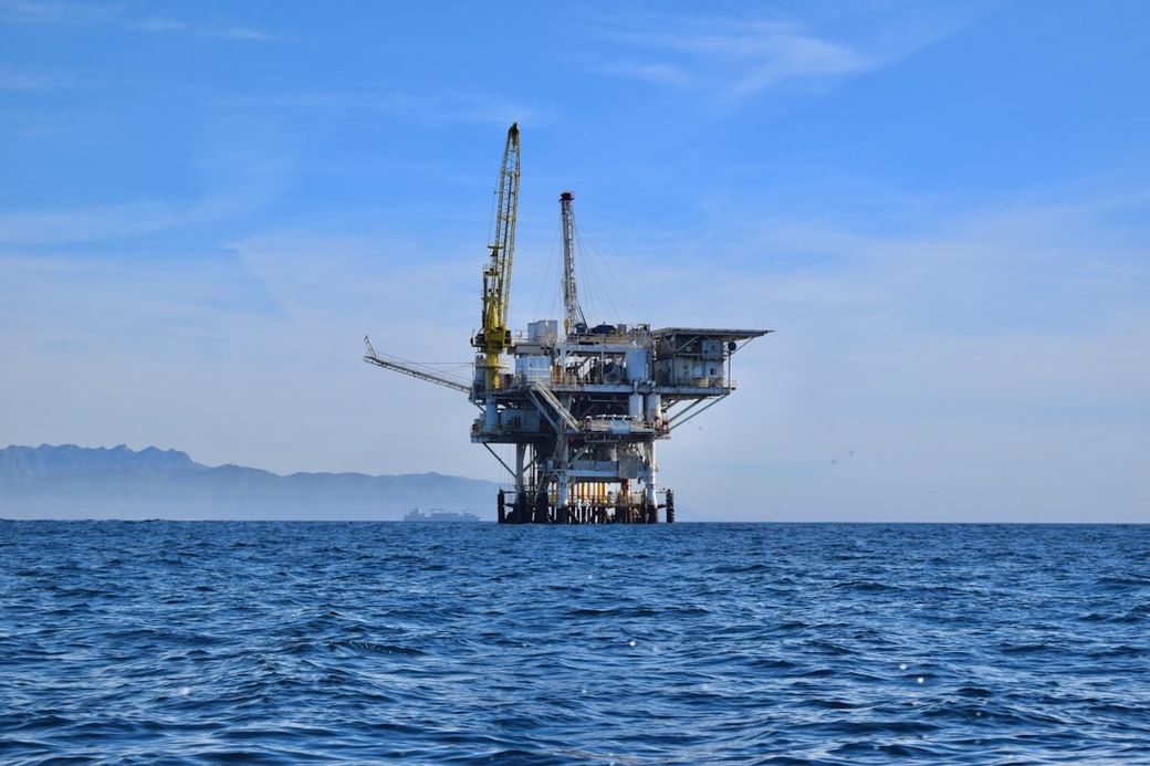 An offshore oil rig stands prominently in the ocean against a clear sky. The structure features cranes and multiple platforms, indicating active extraction operations. — Credit: Photo by Zach Theo on Unsplash