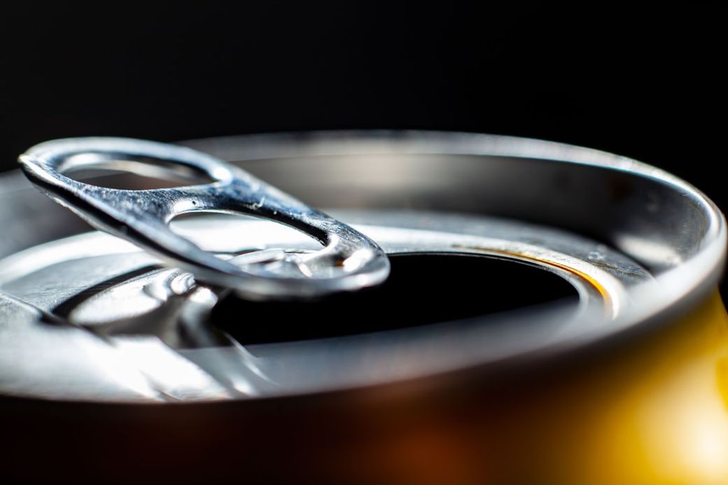 This image features a close-up view of the opening tab on a beverage can. The metallic finish and the circular rim of the can are prominently highlighted against a dark background. — Credit: Photo by Ian Talmacs on Unsplash