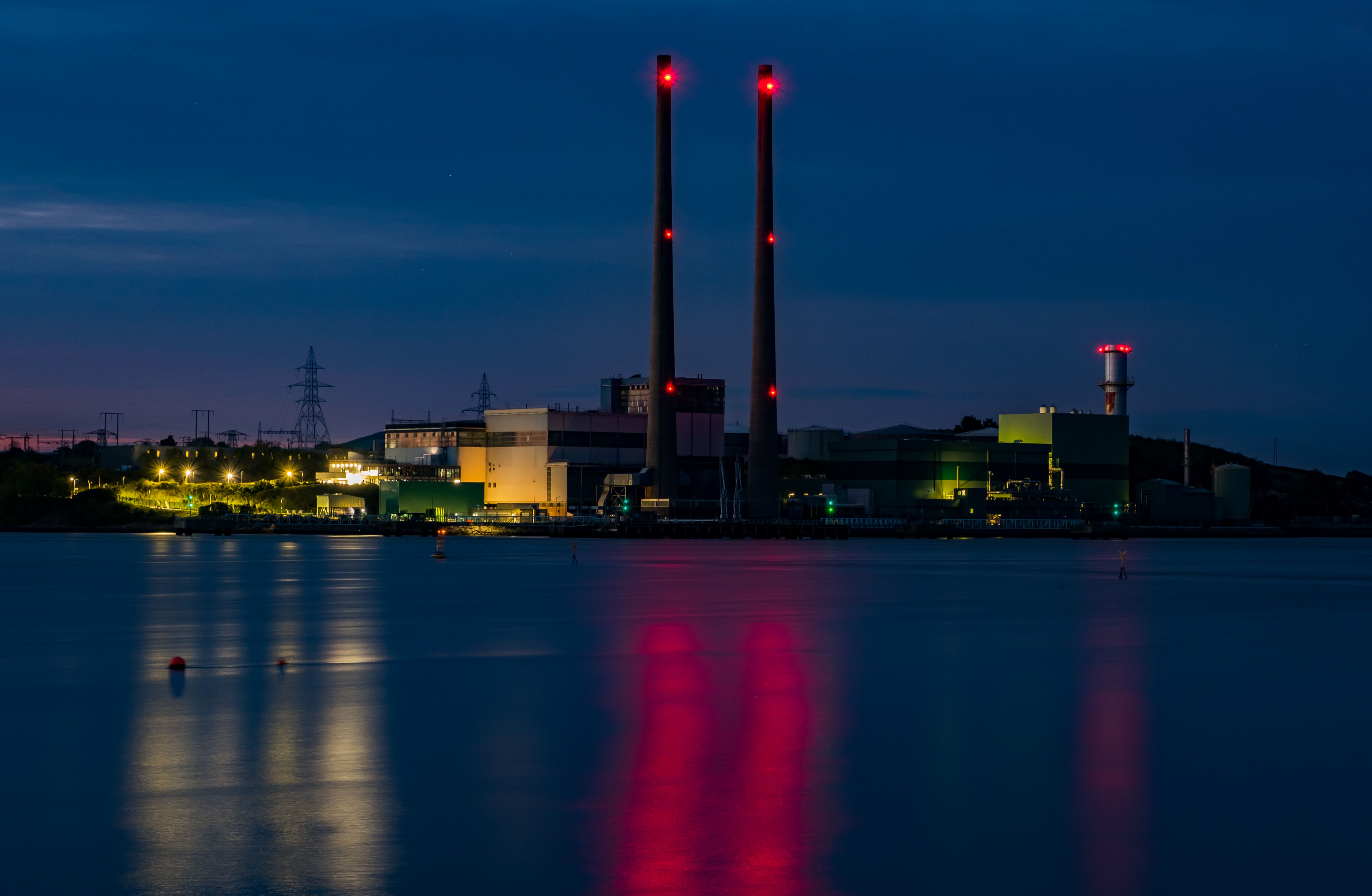 The image showcases a power plant at dusk, highlighted by the glow of industrial lights and the reflection in the calm water below. Two tall smokestacks emit red lights, indicating operational status.