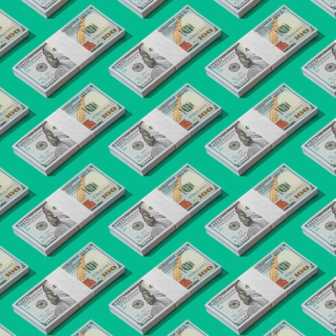 The image features a repeated pattern of stacked bundles of hundred dollar bills against a bright teal background. The arrangement emphasizes the design of U.S. currency, highlighting its intricate details.
