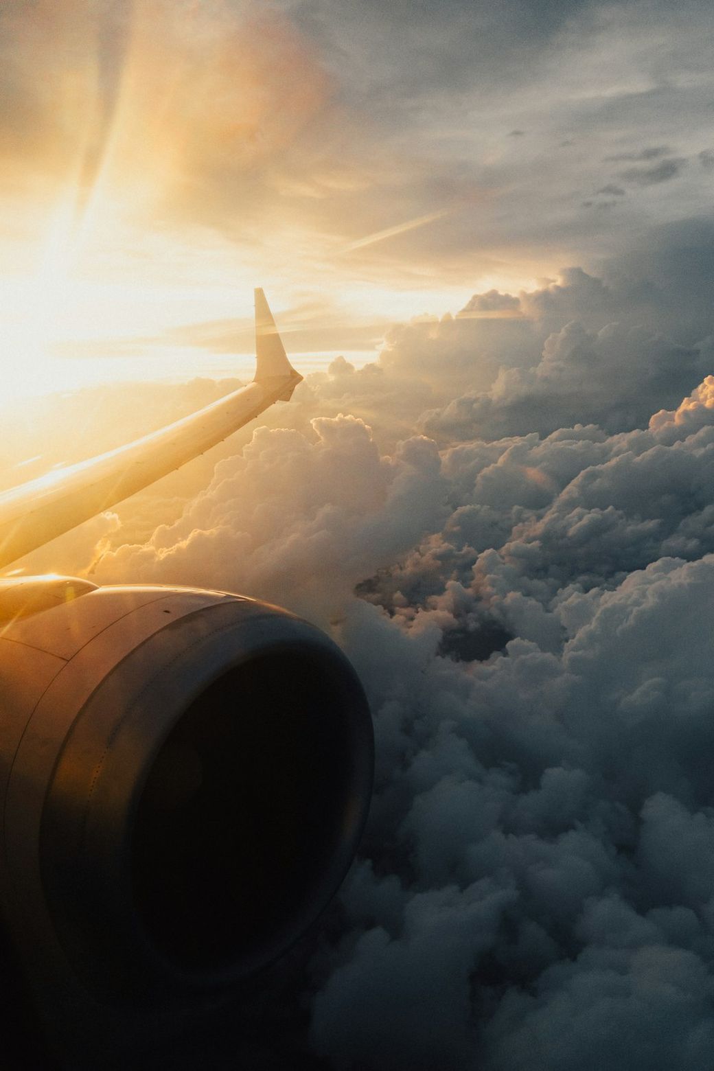 An airplane wing extends out over a dramatic cloudscape, bathed in golden sunset light. The image captures a serene yet awe-inspiring aerial view, showcasing the calmness of flying above the clouds. — Credit: Photo by Tolga Ahmetler on Unsplash