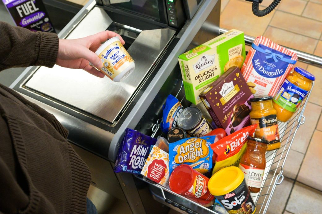 The image shows a person holding a small container of cream while standing next to a checkout counter filled with various grocery items in a shopping basket. The scene captures a typical supermarket environment during checkout.