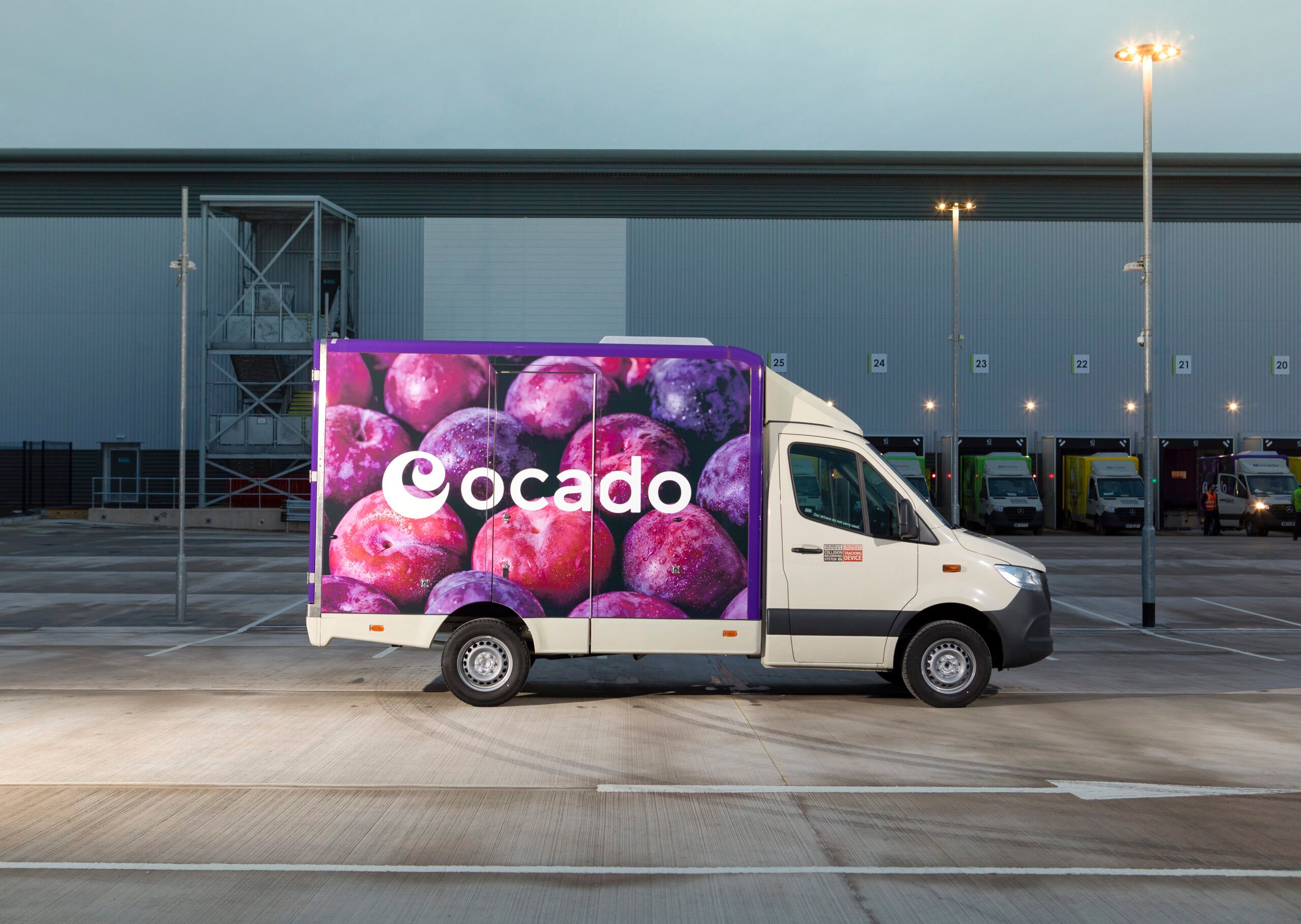 A delivery truck with a vibrant advertisement featuring various fruits is parked outside a warehouse. The setting appears to be an industrial area, possibly related to logistics or food distribution.