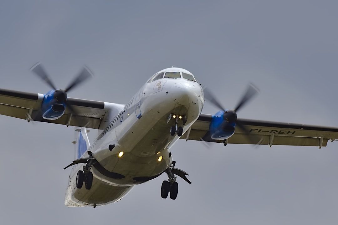 A low-flying propeller aircraft is captured in flight, with its landing gear extended and lights on. The aircraft appears to be approaching a runway against a cloudy sky.