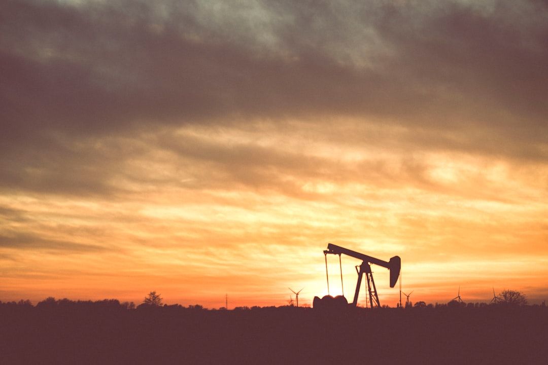 The image shows a silhouette of an oil pump jack against a vibrant sunset sky. Wind turbines are visible in the background, indicating a landscape where traditional and renewable energy sectors intersect.