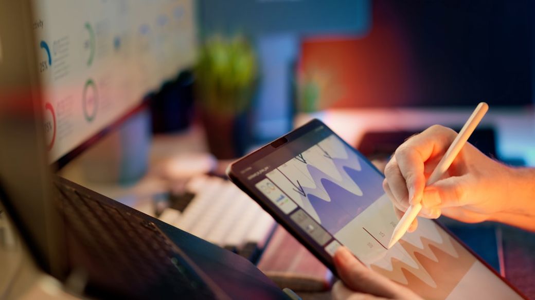A close-up image of a person using a stylus on a tablet device while sitting in front of two computer monitors. The setting appears to be a modern workspace, with vibrant lighting and a hint of greenery in the background. — Credit: Photo by Jakub Żerdzicki on Unsplash