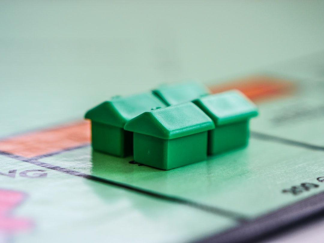 The image features several green plastic houses placed on a game board, suggesting a real estate theme. The board is reminiscent of a property trading game, commonly associated with Monopoly.