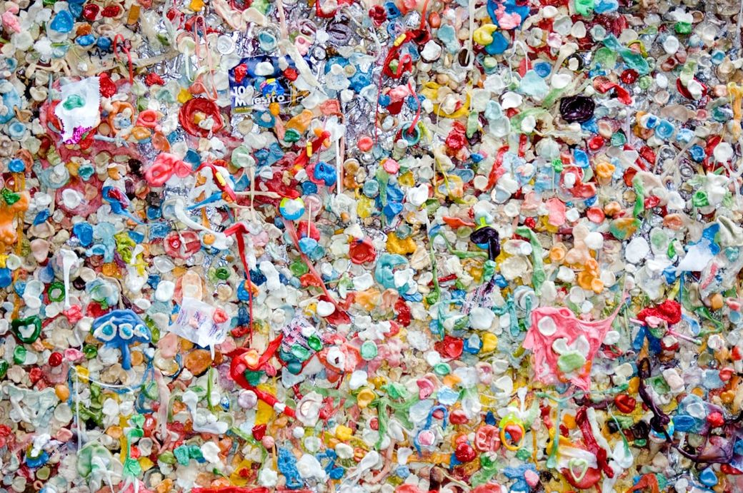 The image depicts a vibrant wall covered in a multitude of colorful chewing gum pieces, creating a chaotic but artistic visual texture. The various hues and textures of the gum create an abstract, almost mosaic-like appearance. — Credit: Photo by Marc Newberry on Unsplash