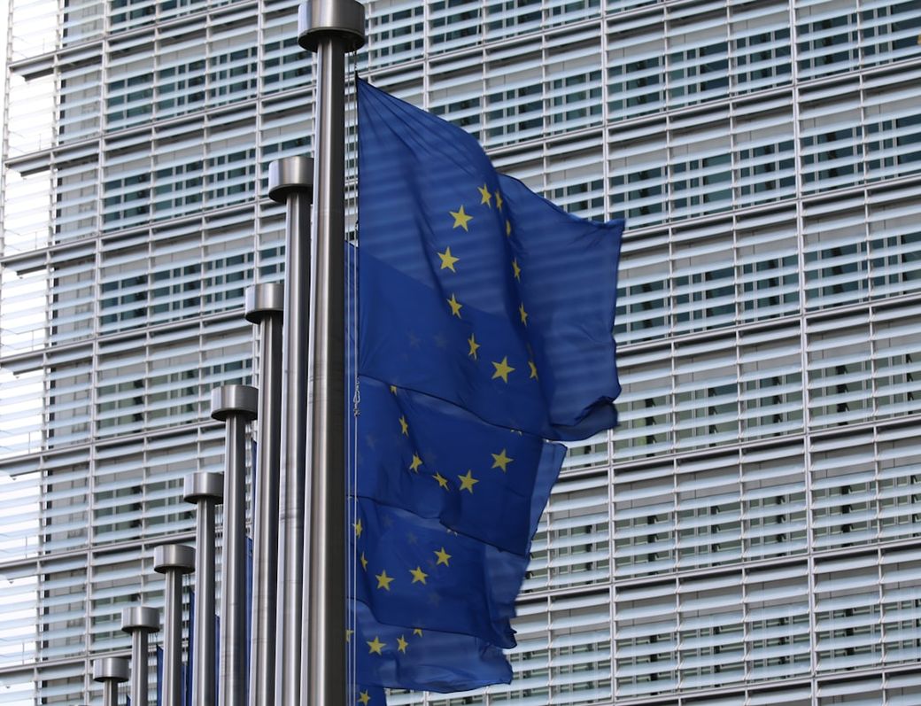 The image depicts several European Union flags waving in front of a modern architectural building. The façade of the building features a grid of windows, typical of contemporary European design. — Credit: Photo by Guillaume Périgois on Unsplash