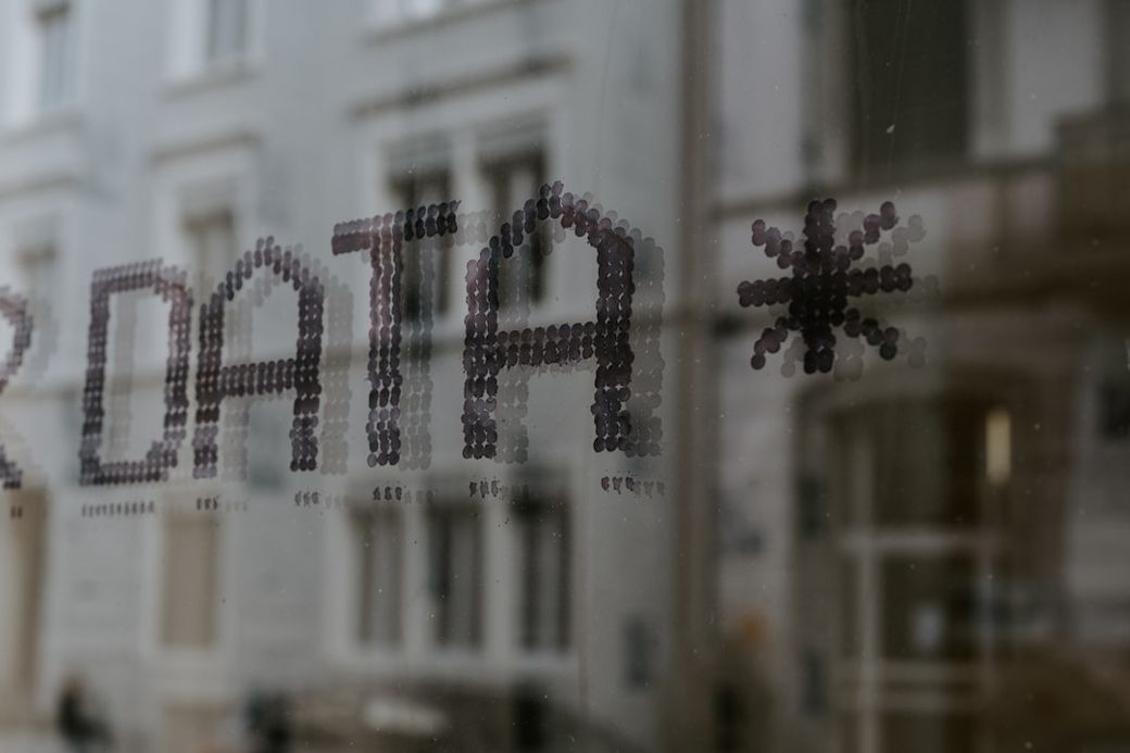 The image depicts a close-up view of a glass window with the word 'DATA' partially visible, reflected against a muted urban background. The letters are formed with small dots, creating a pixelated effect. — Credit: Photo by Claudio Schwarz on Unsplash