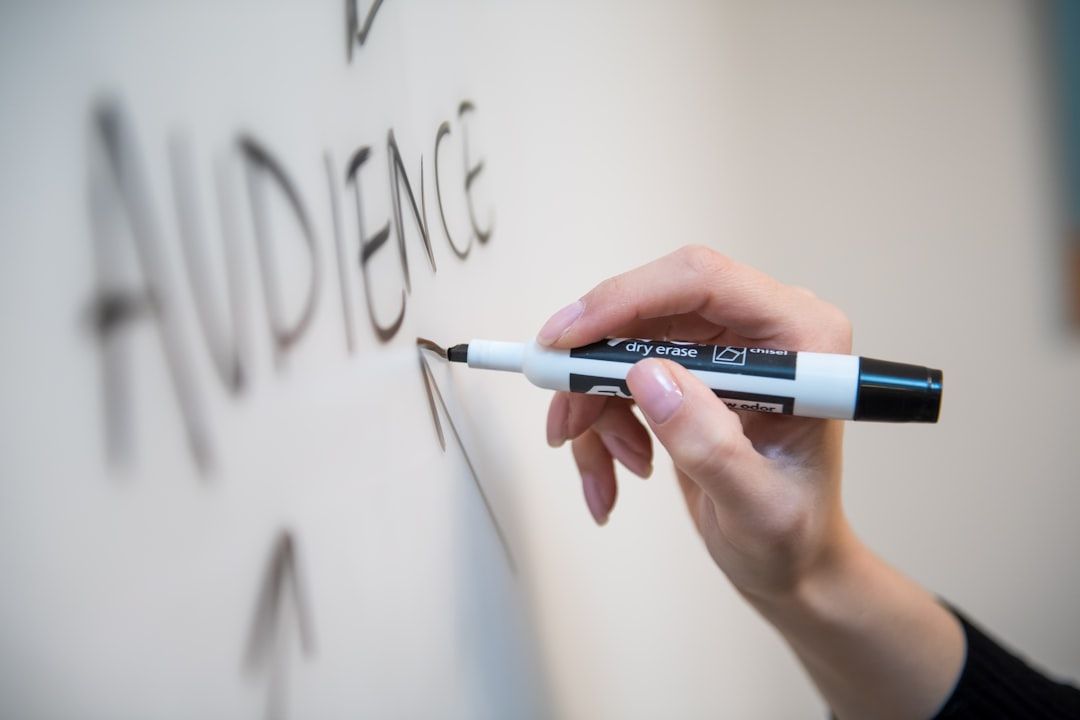 A person is writing the word 'AUDIENCE' on a whiteboard using a dry erase marker. The close-up focus on the hand and marker illustrates the act of preparing for a presentation or discussion.