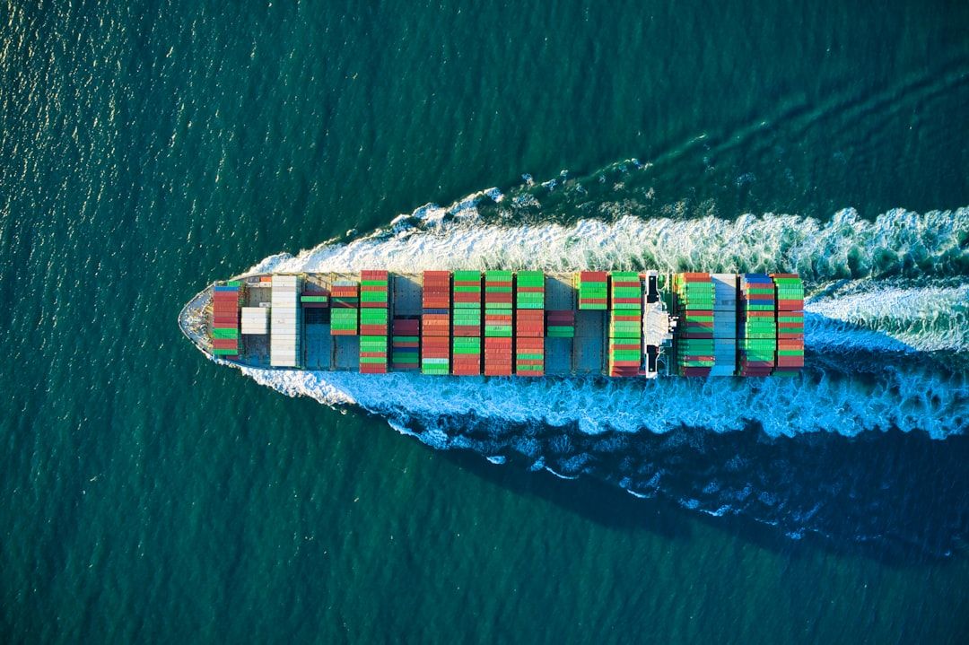 An aerial view of a large cargo ship navigating through open waters. The ship is loaded with colorful shipping containers, creating a vibrant pattern against the deep blue sea.